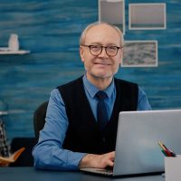 retired-manager-sitting-desk-front-camera-smiling-after-typing-laptop-working-from-home-while-senior-wife-reading-book-background_482257-2442